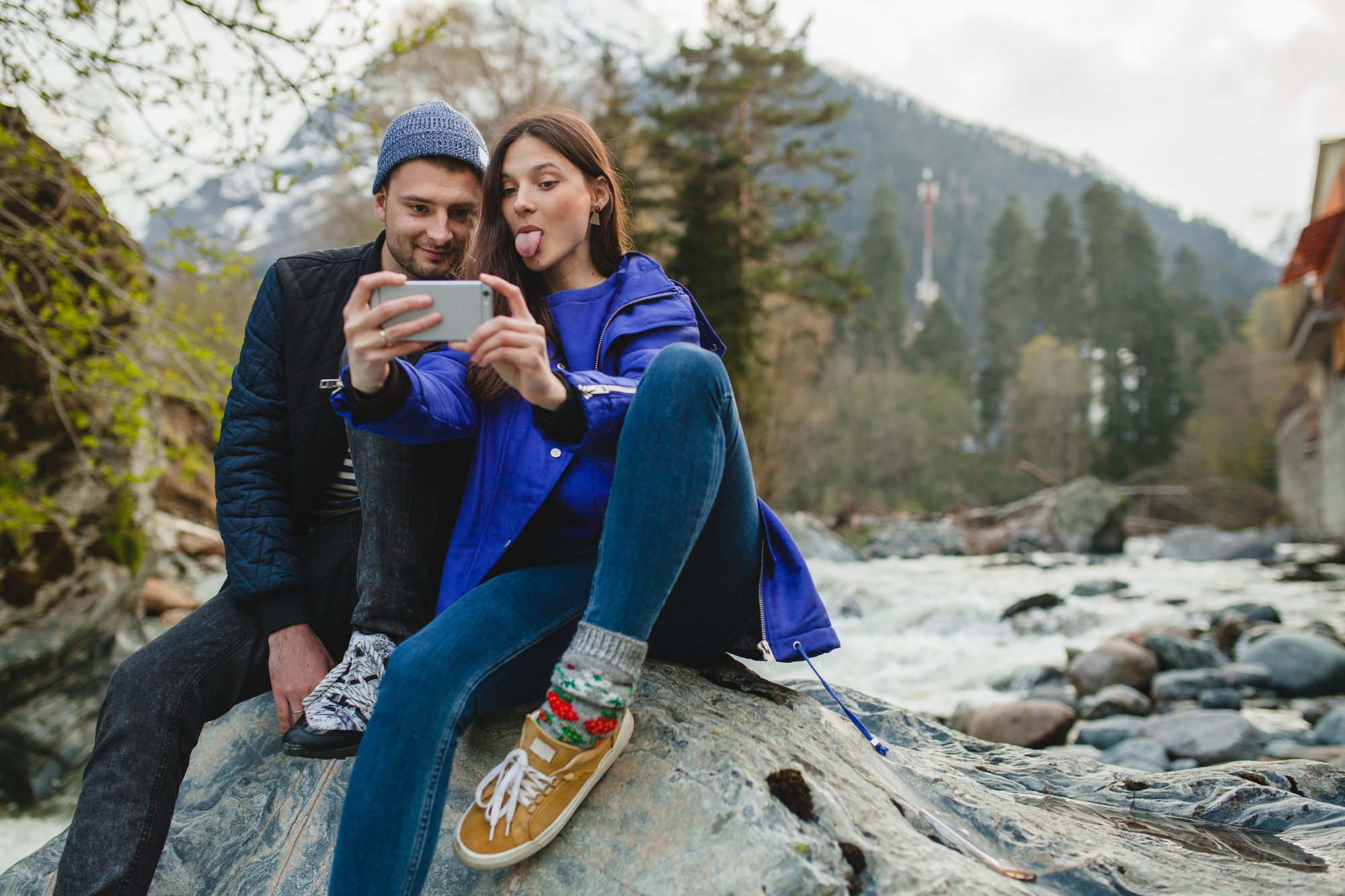 Young couple taking a playful selfie on a creek rock with snow-capped mountains and pine forest behind them