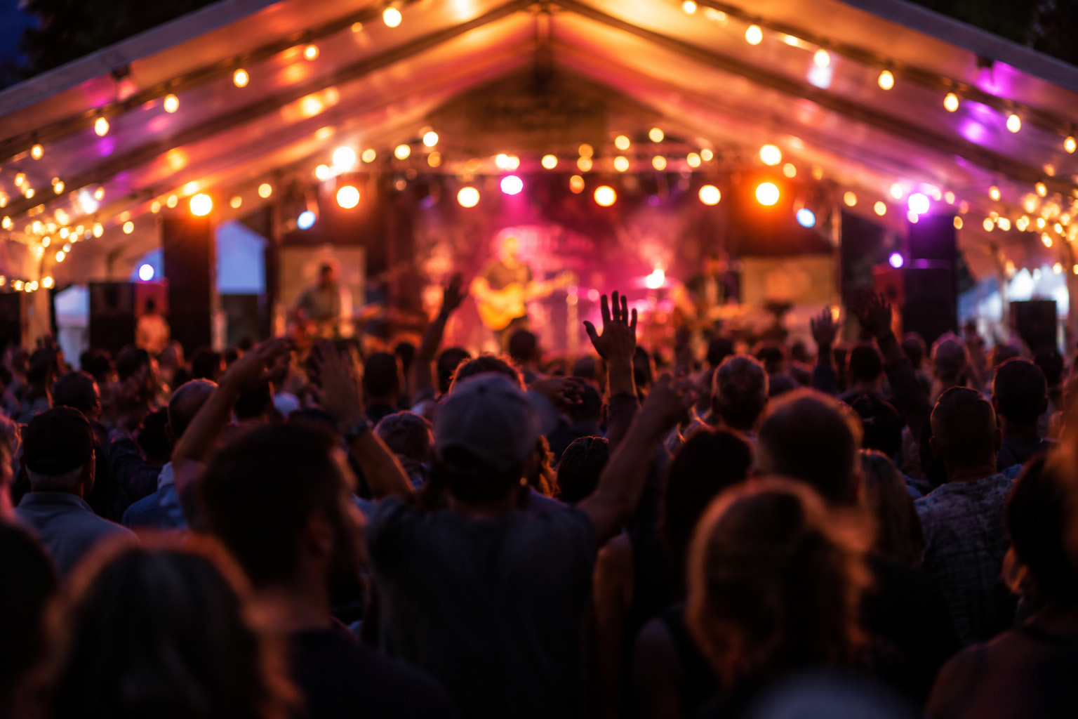A summer mountain concert at twilight, crowd silhouettes with hands raised, café string lights overhead, warm stage glow under a festival tent